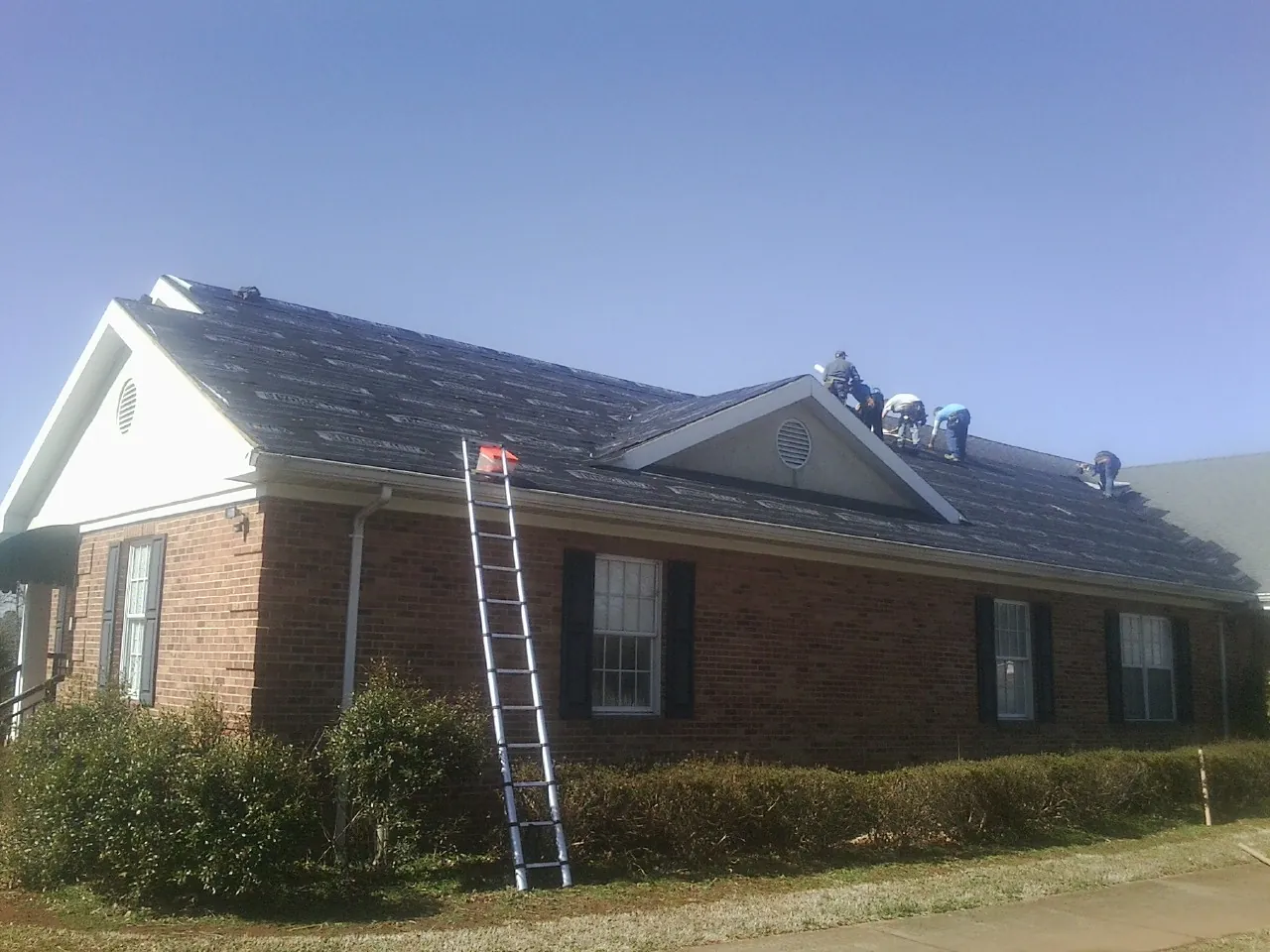 Crew tearing shingles off a brick ranch roof with a ladder set — Emergency Roof Repair work by our crew in Lititz