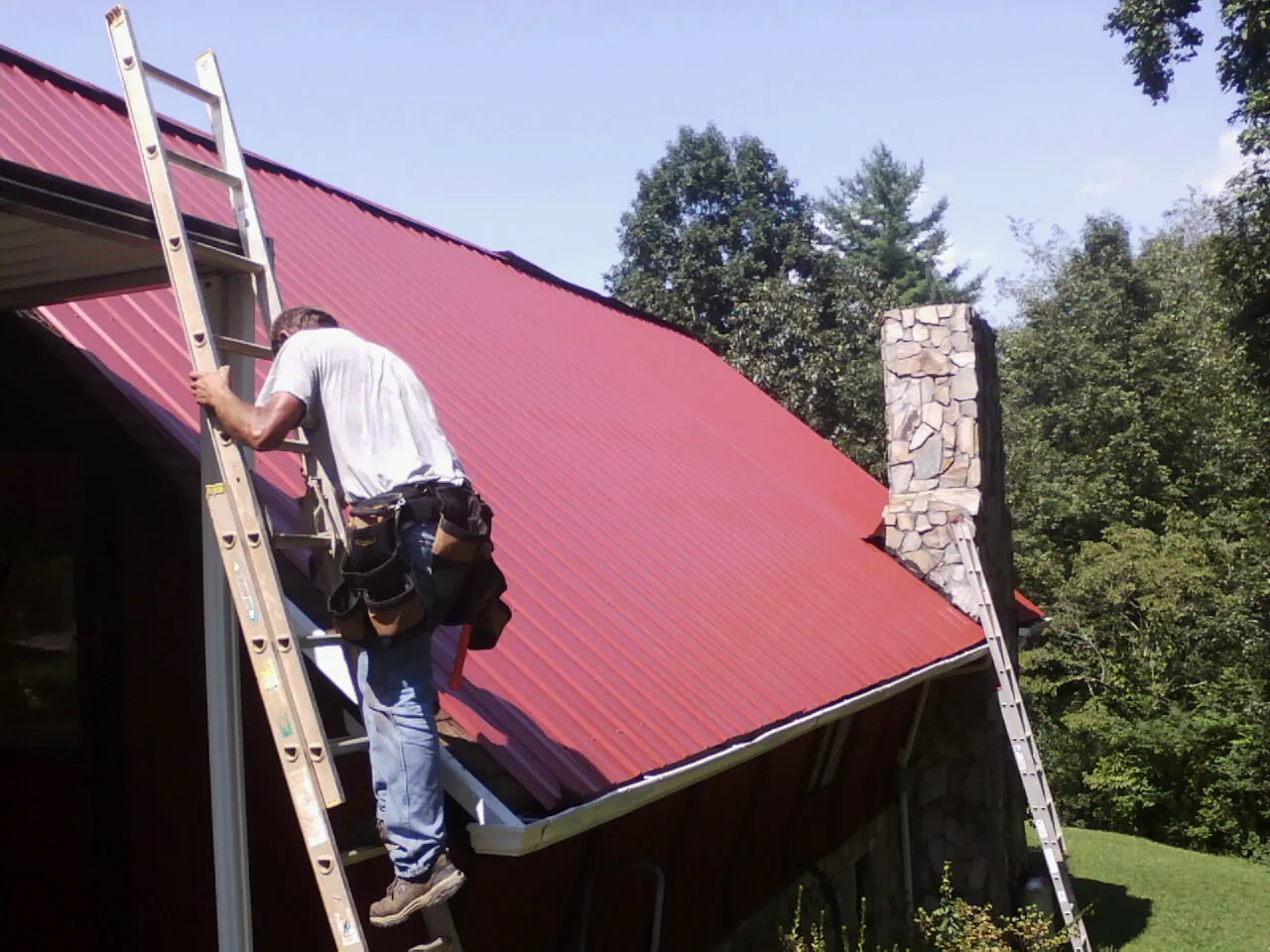 Roofer on a ladder beside a red metal roof and stone chimney — Soffit & Fascia Repair by Roofing Brothers in Lititz